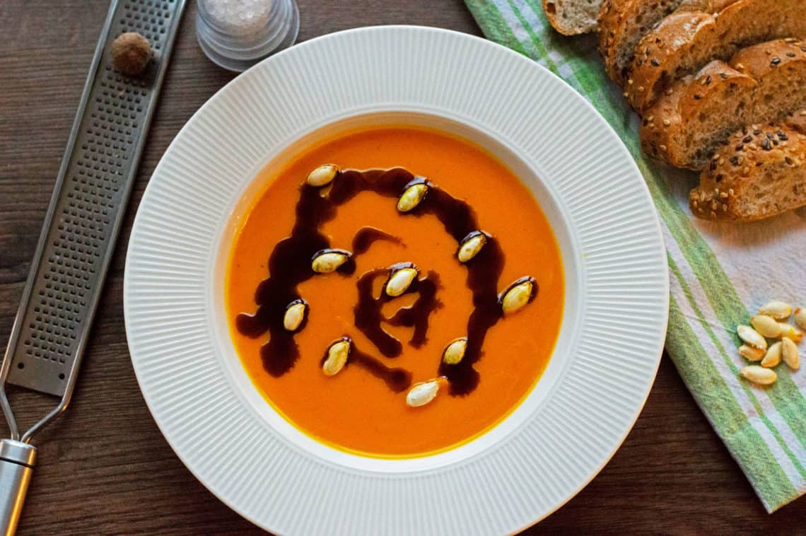 A top shot photo of Pumpkin Soup with nutmeg, pumpkin oil and pumpkin seeds served with some wholegrain bread