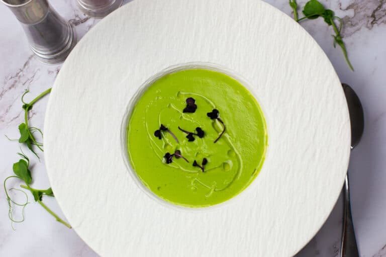 Pea soup with white truffle oil in a white bowl garnished with micro herbs.