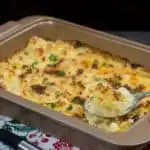 A baking tray with freshly baked cauliflower cheese with a crunchy topping. A spoon taking out a piece of cauliflower from the baking tray.