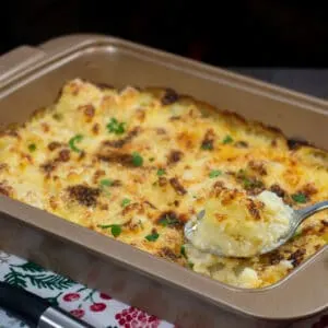 A baking tray with freshly baked cauliflower cheese with a crunchy topping. A spoon taking out a piece of cauliflower from the baking tray.