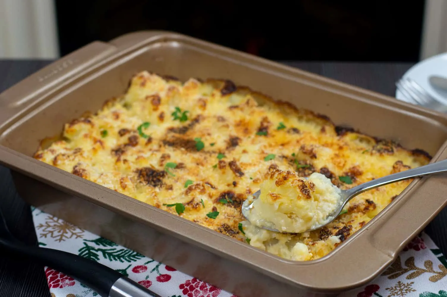 A baking tray with freshly baked cauliflower cheese with a crunchy topping. A spoon taking out a piece of cauliflower from the baking tray.