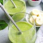 Two glasses of green smoothie with fruit in the background