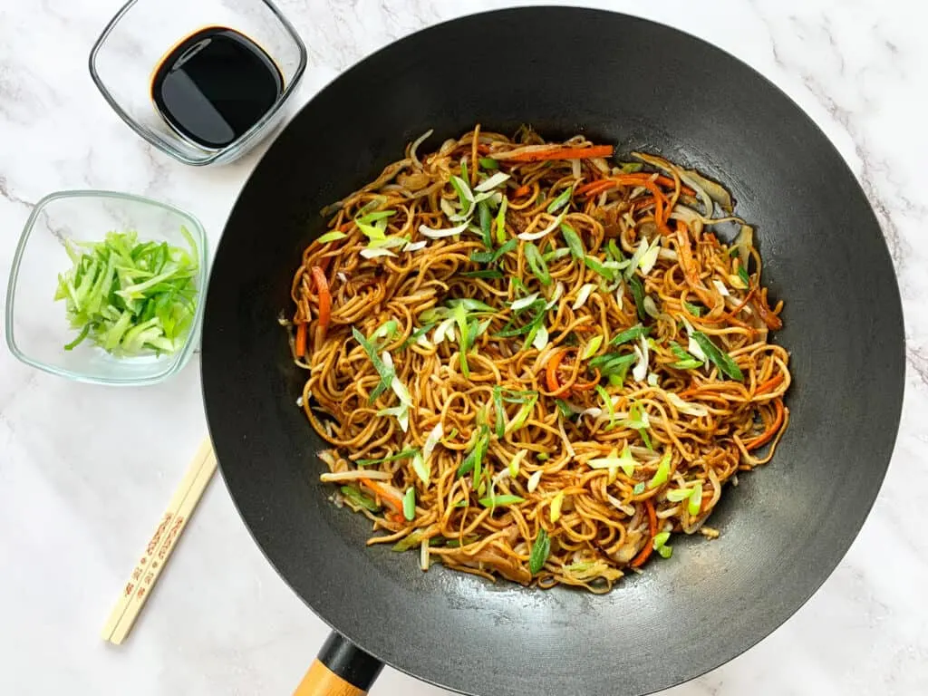 A wok full of vegetable chow mein with soy sauce and spring onions on the side