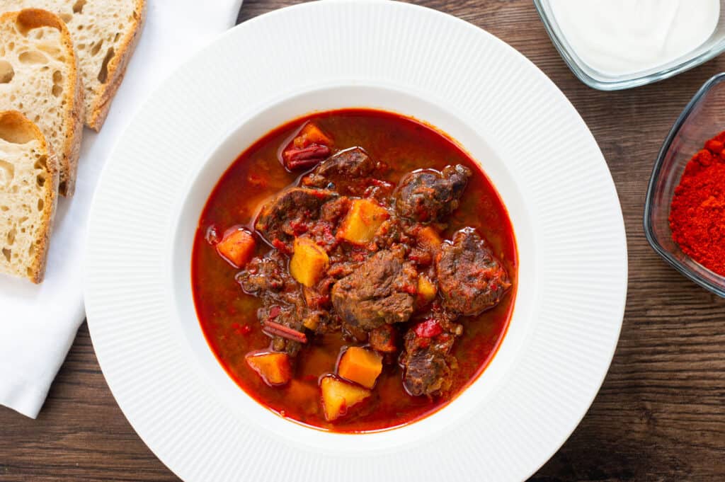 Traditional Hungarian Goulash in a white bowl with sides of sour cream and bread