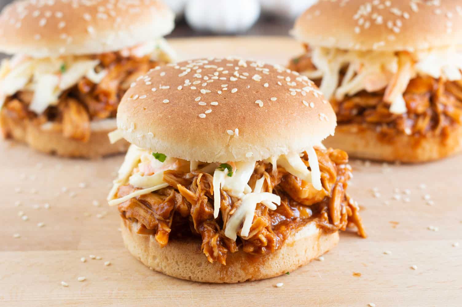 Three pulled pork sandwiches on a wooden surface. Each sandwich is filled with shredded pork in barbecue sauce, topped with coleslaw, and placed in a sesame seed bun.