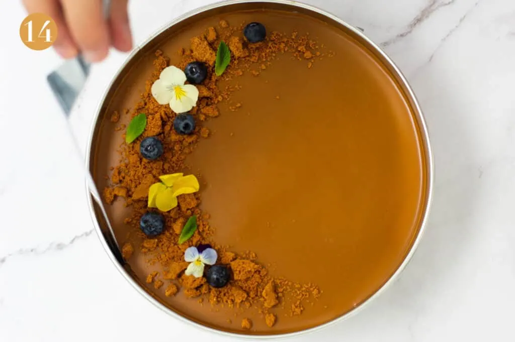 Blueberries, biscoff biscuits, edible flowers and mint on top of the cheesecake in a cake tin with a spatula cutting around the edges
