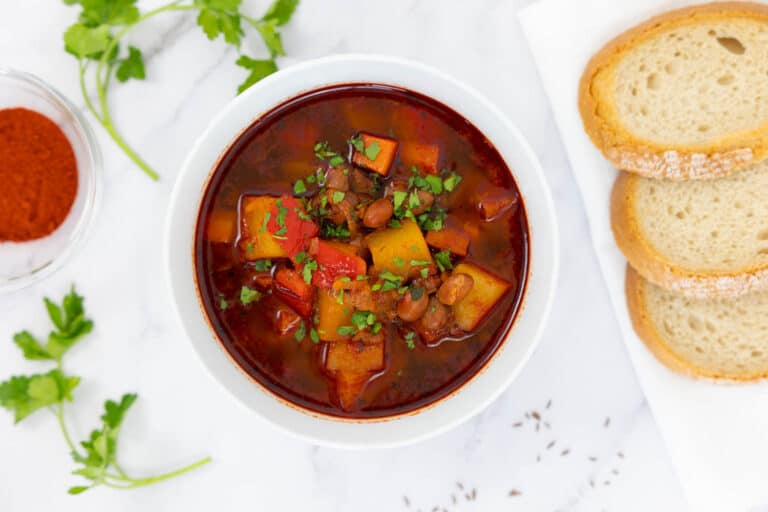 Vegan goulash in a white bowl with Hungarian paprika, parsley and bread beside it