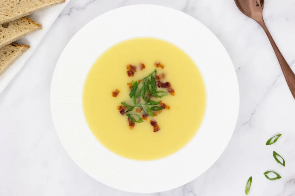Potato soup in a fancy white bowl topped with crispy bacon and sliced spring onions with a side of bread