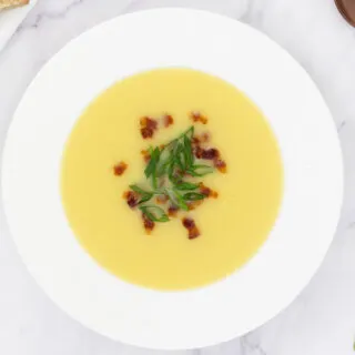 Potato soup in a fancy white bowl topped with crispy bacon and sliced spring onions with a side of bread