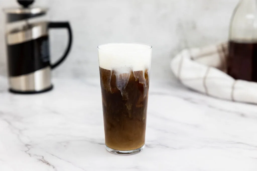 Cold foam on top of a iced cold brew in a glass with a french press and bottle of vanilla syrup in the background