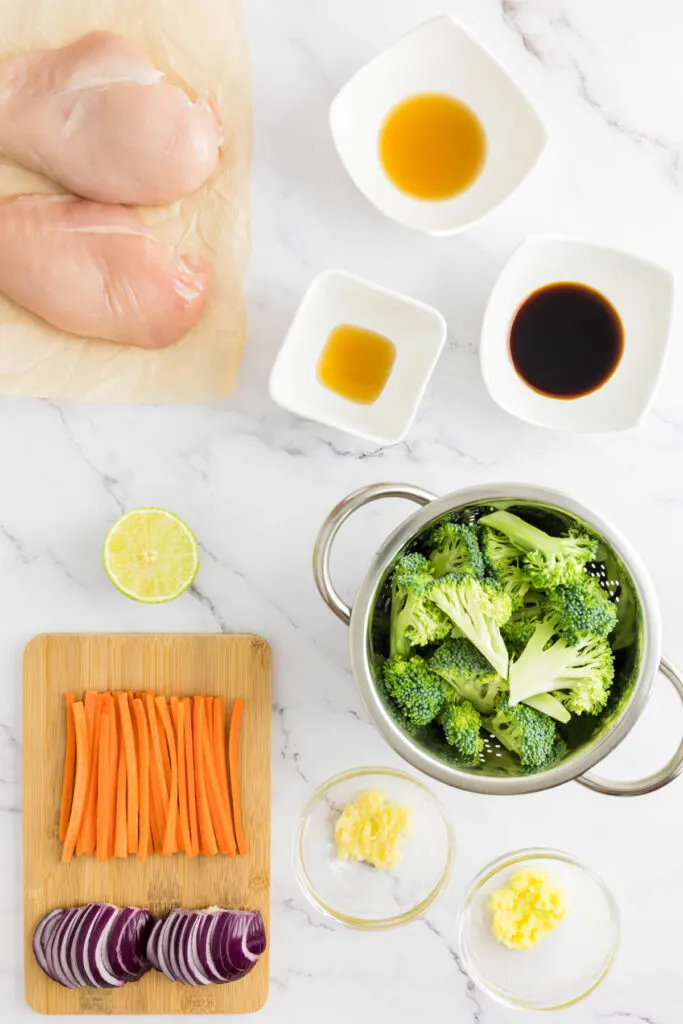 Keto chicken stir fry: Skinless, boneless chicken breasts, soy sauce, vegetable oil, garlic, ginger, carrots, red onion, broccoli, sesame oil, fish sauce & lime portioned on a marble counter