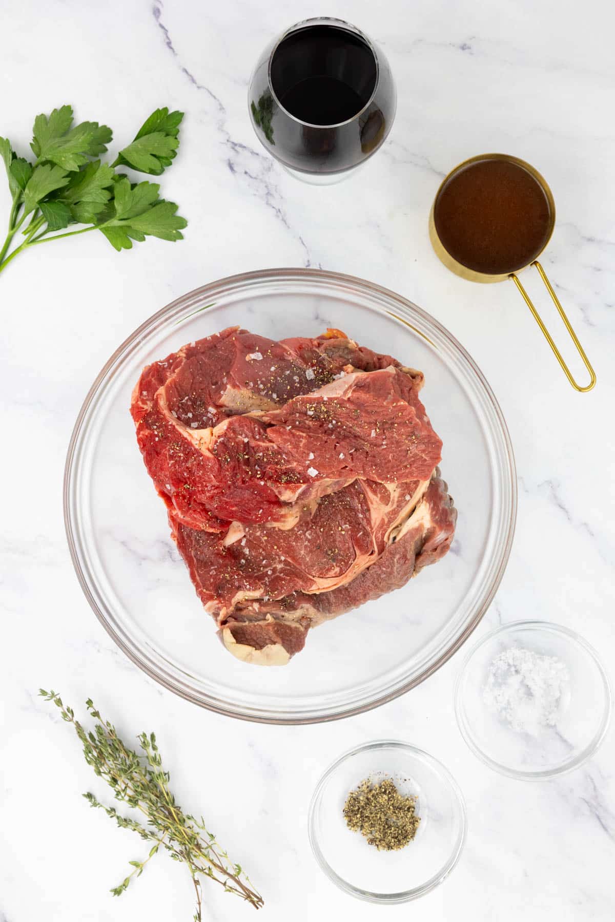 Beef, beef stock, red wine, parsley, thyme, salt and pepper on a table.